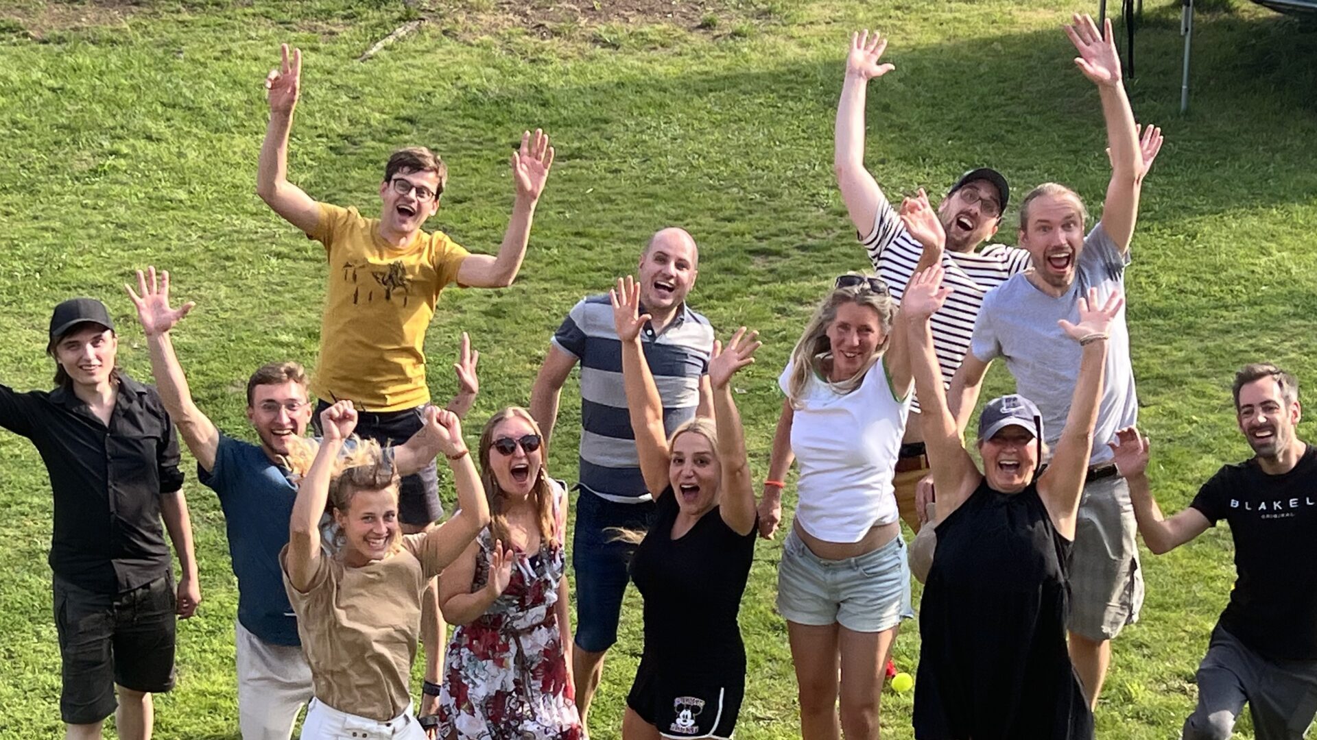 die Hilfswerft Teammitglieder springen synchron in die Höhe, werfen die Arme hoch und lachen. Sie stehen auf einer Wiese und werden von schräg oben fotografiert.