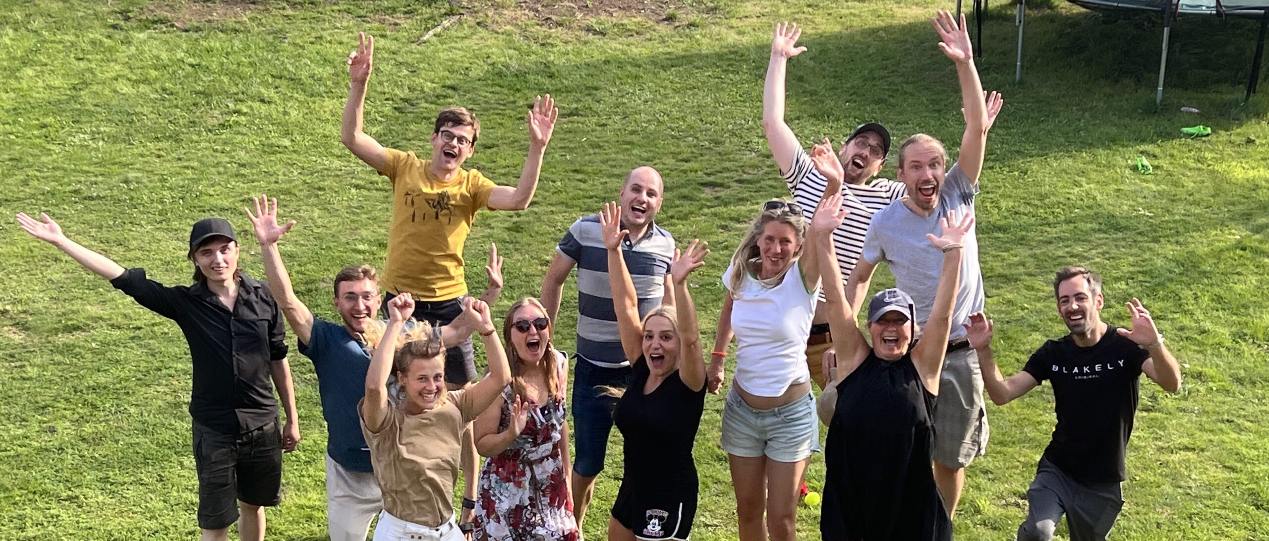 die Hilfswerft Teammitglieder springen synchron in die Höhe, werfen die Arme hoch und lachen. Sie stehen auf einer Wiese und werden von schräg oben fotografiert.